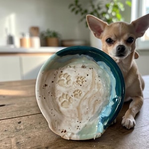 May include: A round ceramic spoon rest featuring a speckled cream-colored center with embossed paw prints and a deep teal blue glazed rim. The surface has a smooth, glossy finish with subtle variations in color and texture throughout the piece.