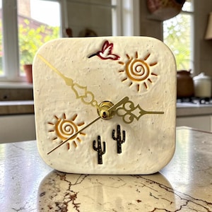 Puede incluir: Un reloj cuadrado de color crema con un tema desértico. La esfera del reloj presenta dos soles, dos cactus y un colibrí rojo. Las manecillas del reloj son doradas. El reloj está sobre una encimera de mármol.