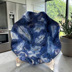 May include: A decorative, blue leaf-shaped bowl with a glossy finish. The bowl features a detailed leaf pattern in a lighter blue hue, creating a contrast. It is displayed on a clear stand, showcasing its unique design. Ideal for home decor.