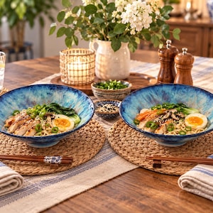 May include: Two deep, round ceramic bowls with a vibrant blue, mottled glaze containing ramen noodles, sliced chicken, soft-boiled eggs, bok choy, and green onions. Each bowl is set on a woven placemat with a pair of wooden chopsticks resting nearby.