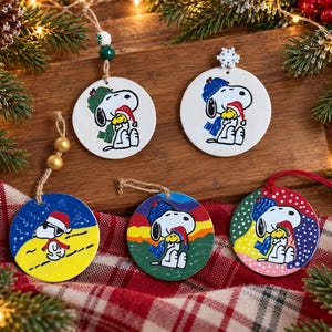 May include: Five round ceramic ornaments featuring Snoopy and Woodstock. Two white ornaments show the characters in winter hats. Three colorful ornaments depict Snoopy in a Santa hat on a yellow background, a sunset landscape, and a polka-dot pattern.