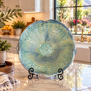 May include: A round ceramic bowl featuring a swirling blue and yellow glazed finish with a prominent spiral design at the center. The bowl has a scalloped edge and is displayed upright on a black metal stand, showcasing its textured surface.