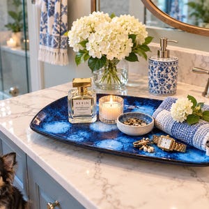 May include: A rectangular ceramic tray with a deep blue and white mottled glaze. The tray holds a square glass perfume bottle labeled AERIN, a small lit candle in a glass holder, a stone bowl with jewelry, a gold wristwatch, and a blue and white towel.