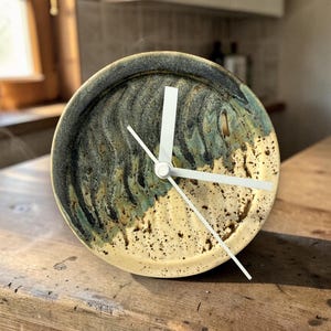 May include: A ceramic clock with a cream-colored face and a dark blue and green rim. The clock hands are white. The clock is sitting on a wooden surface. The clock face has a speckled design.