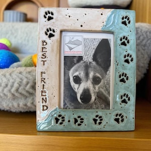 May include: Ceramic photo frame with a picture of a dog. The frame is rectangular with a light beige and blue speckled finish. Black paw prints and the words "BEST FRIEND" are on the frame.