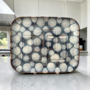 May include: A rectangular decorative tray with a unique pattern of white and blue circles. The tray has a bronze-colored rim and is placed on a white countertop. The design gives the tray a textured appearance.
