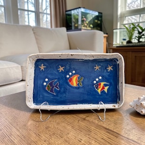 May include: A rectangular ceramic tray with a blue interior, featuring three colorful fish and starfish designs. The tray is supported by a wire stand and sits on a wooden table. A white seashell is next to the tray.