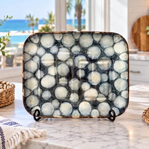 May include: A rectangular ceramic serving platter featuring a reactive glaze finish with a pattern of overlapping dark blue and cream-colored circles. The platter has rounded corners and a dark brown rim, displayed upright on a black metal stand.