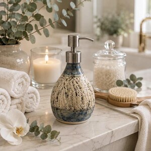 May include: A ceramic soap dispenser with a teardrop shape, featuring a textured cream-colored upper section with dark speckles and a smooth, deep blue glazed base. The dispenser is topped with a brushed silver-tone metal pump mechanism.
