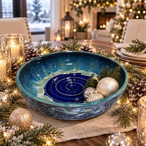 May include: A large, round ceramic bowl featuring a deep blue center with a spiral pattern, transitioning into shades of teal and green toward the rim. The bowl has a glossy finish and contains several silver and white decorative ornaments.