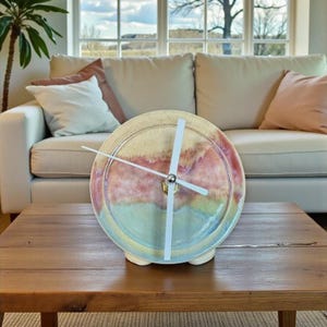 May include: A ceramic clock with a colorful, abstract design in shades of beige, pink, and blue. The clock has white hands and a silver center, resting on a wooden table. The clock's diameter is approximately 8 inches.