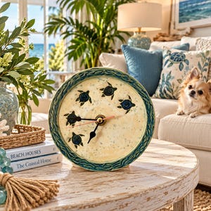May include: A round ceramic clock with a cream-colored face featuring five small dark blue sea turtle illustrations. The clock has a textured teal border with a braided pattern and black clock hands. It sits upright on a round wooden table.