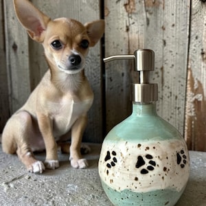 May include: A light brown Chihuahua dog sits next to a ceramic soap dispenser. The dispenser is round with a light green and white color scheme, featuring black paw prints. The dispenser has a silver pump.