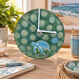 May include: A round ceramic wall clock featuring a mottled green glaze with a circular bubble pattern. A stylized blue sea turtle and a small blue spiral are embossed on the lower section. The clock has simple white hands and a clear plastic stand.