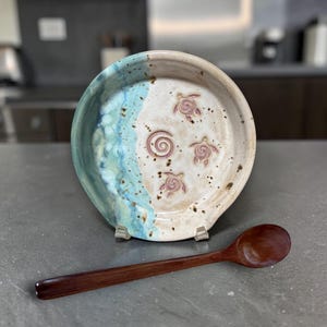 May include: A handmade ceramic spoon rest with a blue and white glaze, featuring turtle designs and a spiral. A dark wooden spoon rests beside the spoon rest. The spoon rest is a kitchen accessory.
