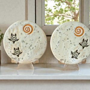 May include: Two decorative ceramic plates with a speckled, off-white finish. Each plate features a black turtle design and an orange spiral. The plates are displayed on clear stands, with a small plant in a glass vase to the left.