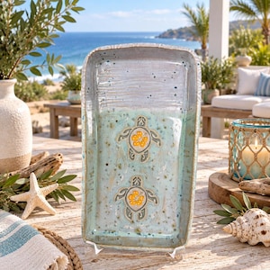 May include: A rectangular ceramic tray with a glossy finish, featuring a two-tone color scheme of light gray at the top and speckled seafoam green at the bottom. Two sea turtle motifs are centered, each with a yellow hibiscus flower shell.