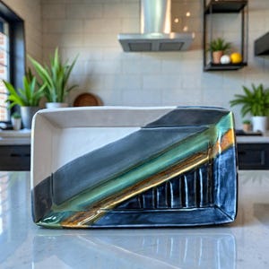 May include: Rectangular ceramic serving dish with a modern design. The dish features a white interior, a diagonal section of dark gray, green, and gold, and a dark blue exterior. The dish is displayed on a white countertop.