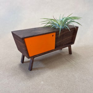 May include: A miniature mid-century modern style cabinet with a single orange door and a planter on top. The cabinet is made of dark wood and has four legs.