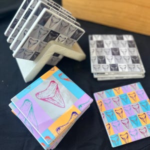 May include: A set of square ceramic coasters with shark tooth designs. Some coasters feature a black and white pattern, while others showcase colorful designs with pink, blue, orange, and purple hues. The coasters are displayed in a white holder.