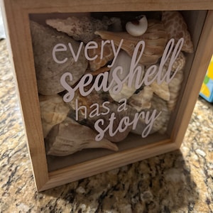 May include: A wooden shadow box with a glass front filled with seashells. The box has a white inscription that reads "every seashell has a story".