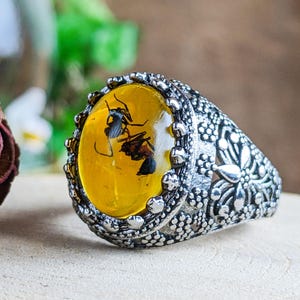 May include: A silver ring with an oval amber stone, showcasing preserved insects. The amber is a vibrant yellow, set in a detailed, floral-patterned silver band. The ring is displayed on a wooden surface.