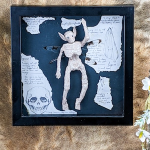 May include: A framed shadow box art piece featuring a skeletal figure with bat-like wings, set against a dark background with torn paper and sketches. The artwork includes a skull drawing and handwritten notes, all within a black frame.