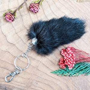 May include: A black fur keychain with a silver clasp and ring. The keychain is on a wooden surface, with dried pink flowers and red and green decorative sprigs.