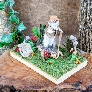 May include: A whimsical diorama featuring a white, furry creature in a cowboy hat and vest, holding a walking stick. The scene is set on a wooden base covered in green moss, with miniature plants and a log. The background is a textured brown.