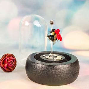 May include: A taxidermy bumblebee with a bright red bow attached to its back, mounted on a clear vertical pin atop a circular black wooden base. The display includes miniature paper currency and is covered by a removable clear glass dome.