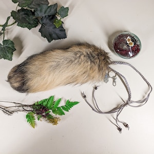 May include: A fluffy brown and gray faux fur tail with a gray braided cord and dark brown beads. The tail is attached to the cord with a silver charm.