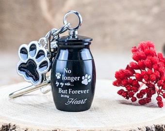May include: Black pet memorial urn keychain with a paw print charm. The urn has the text "No longer by my side... But Forever in my Heart". The keychain is on a wooden surface with red flowers.