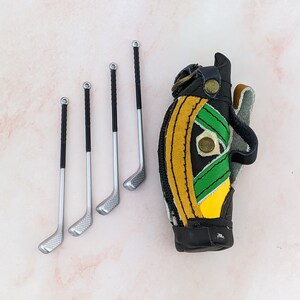 May include: Miniature golf club set with four clubs and a golf bag. The clubs have silver heads and black handles. The golf bag is black with yellow and green accents. The set is likely a decorative item or a toy.