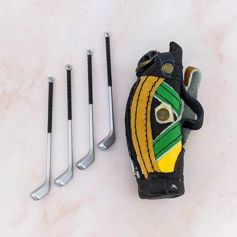 May include: Miniature golf club set with four clubs and a golf bag. The clubs have silver heads and black handles. The golf bag is black with yellow and green accents. The set is likely a decorative item or a toy.