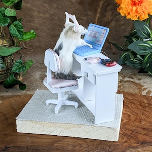 May include: A miniature scene featuring a small, furry creature wearing headphones and seated at a tiny white desk. The creature is using a blue laptop, with a pink chair and other accessories. The scene is set on a textured base.