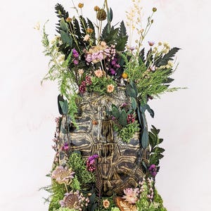 May include: A decorative, textured, brown and black turtle shell sculpture, adorned with a variety of dried flowers, greenery, and moss. The arrangement includes purple, pink, and white flowers, creating a natural, earthy aesthetic. The sculpture is set on a wooden base.
