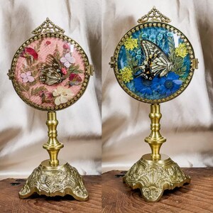 May include: Two decorative butterfly displays. Each display features a round, glass-covered frame with a butterfly and dried flowers. The frames are set on ornate, gold-colored metal stands. One display has a pink background, the other blue.