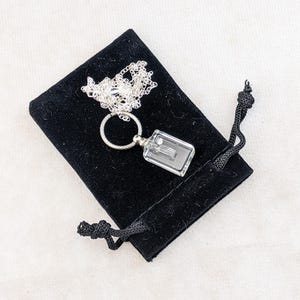 May include: A silver necklace with a rectangular, clear pendant and a delicate chain, resting on a black velvet pouch. The pendant has a small, detailed design inside. The pouch is closed with a black drawstring.