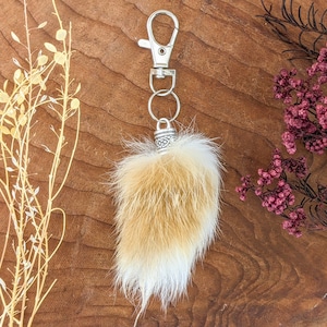 May include: A fluffy keychain charm with a silver-colored clasp and ring. The charm is made of tan and white fur, with a decorative silver-colored cap. The background is a wooden surface with dried plants and flowers.
