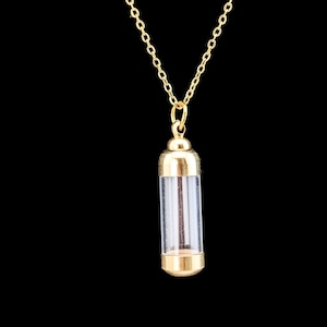 May include: A gold-toned pendant necklace featuring a clear, cylindrical glass vial suspended from a delicate gold-toned chain. The vial is capped with gold-toned metal at both the top and bottom, creating a simple, vertical, tube-shaped design.