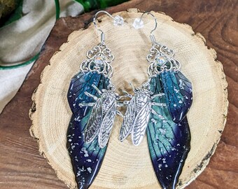 May include: A pair of handcrafted earrings featuring a silver cicada design with blue and green iridescent wings. The earrings have a decorative silver top with a clear crystal accent and a fish hook closure.