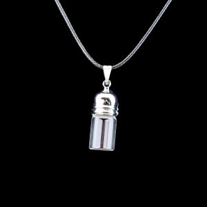 May include: A silver-toned pendant necklace featuring a small, clear glass vial with a metallic cap. The vial hangs from a delicate silver chain against a black background. The pendant is designed to hold small items.