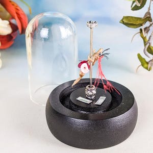 May include: A whimsical sculpture featuring a small creature clinging to a silver pole, adorned with red feathers and a decorative skirt. The sculpture sits on a black wooden base, partially enclosed by a glass dome.