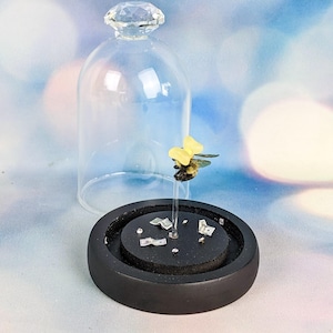 May include: A decorative miniature bee sculpture with yellow wings and a black body, mounted on a clear stand. The bee is displayed inside a glass cloche with a crystal-like top, set on a black base with decorative accents.