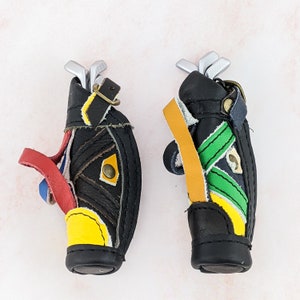 May include: Two miniature golf bag keychains. The bags are black with colorful accents of red, yellow, blue, and green. Each bag has a set of miniature silver golf clubs. The keychains are made of leather and have a strap.