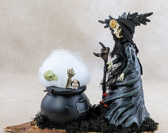May include: A Halloween-themed figurine featuring a witch-like figure with a skull head, black dress, and staff. A black cauldron with a skull, hand, and other items sits on a wooden base. White cotton-like material is in the cauldron.