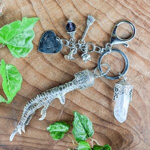 May include: A silver-toned keychain featuring a witch-themed design. Charms include a heart with "WITCH" engraved, a broom, a crystal, and a bird's foot. The keychain is attached to a large ring and a clasp.