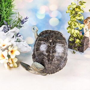 May include: A decorative turtle figurine with a textured, dark brown shell and light green flippers. The figurine has a key-shaped handle and is surrounded by artificial flowers and greenery, creating a whimsical, aquatic theme.