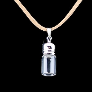 May include: A clear glass vial pendant with a silver-colored cap and bail, suspended from a tan cord. The vial is empty, and the background is black. The pendant is designed to hold small items.