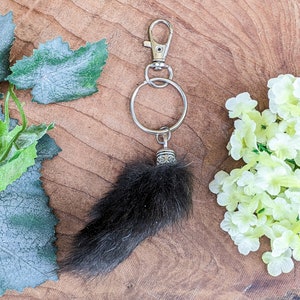 May include: A black fur keychain with a silver-colored clasp and ring. The keychain has a decorative silver-colored cap. The background features green leaves and white flowers on a wooden surface.